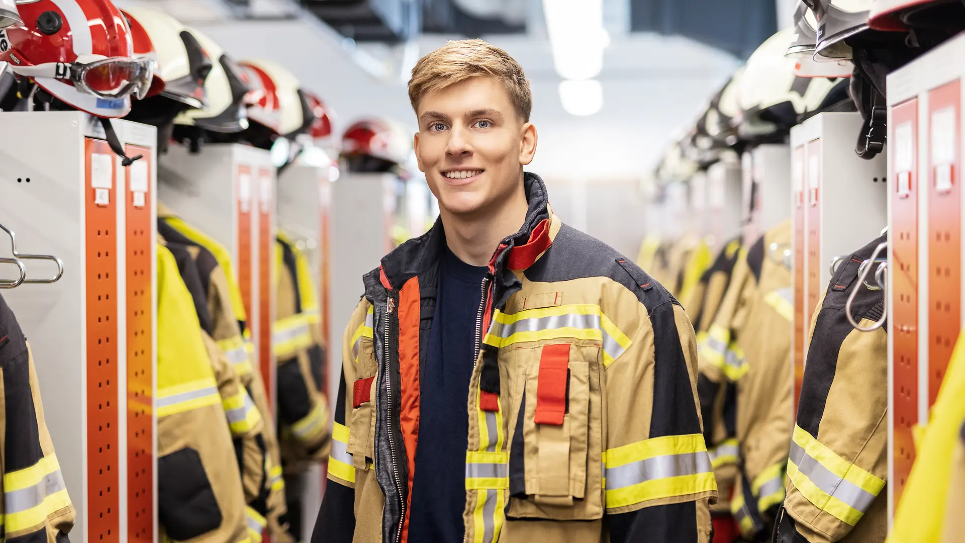 A photo of a smiling young man wearing a fire department uniform and standing in a fire department changing room.