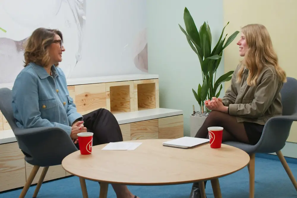 Sylvie and Leonie are sitting opposite each other in a modern, bright room. Both are seated on grey chairs at a round wooden table. On the table, there are two red coffee cups. In the background, there is a plant and a shelf with open compartments.