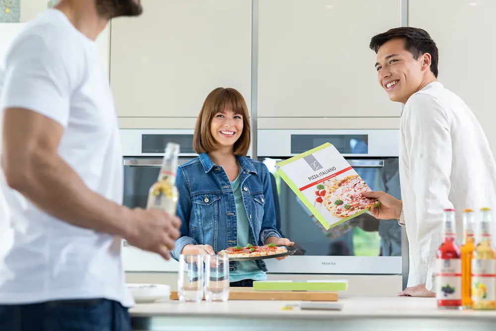
Three people in a kitchen: one holding a bottle, another with a plate of food, and the third with a cookbook, with bottles visible on the counter.