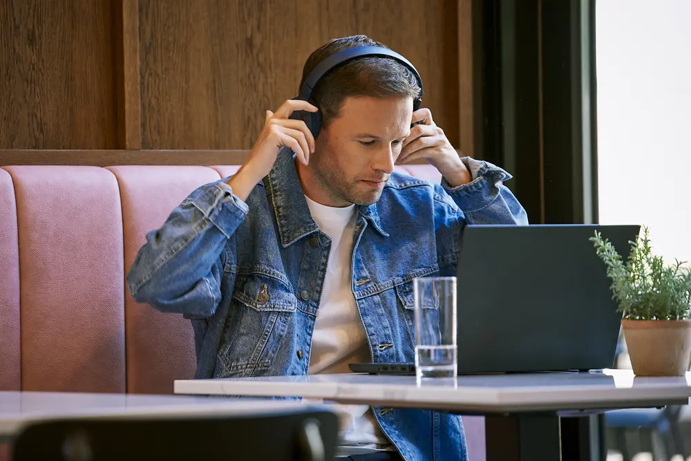 A man wearing a denim jacket and headphones is sitting at a table in a cafe, working on a laptop. There is a glass of water and a potted plant on the table.