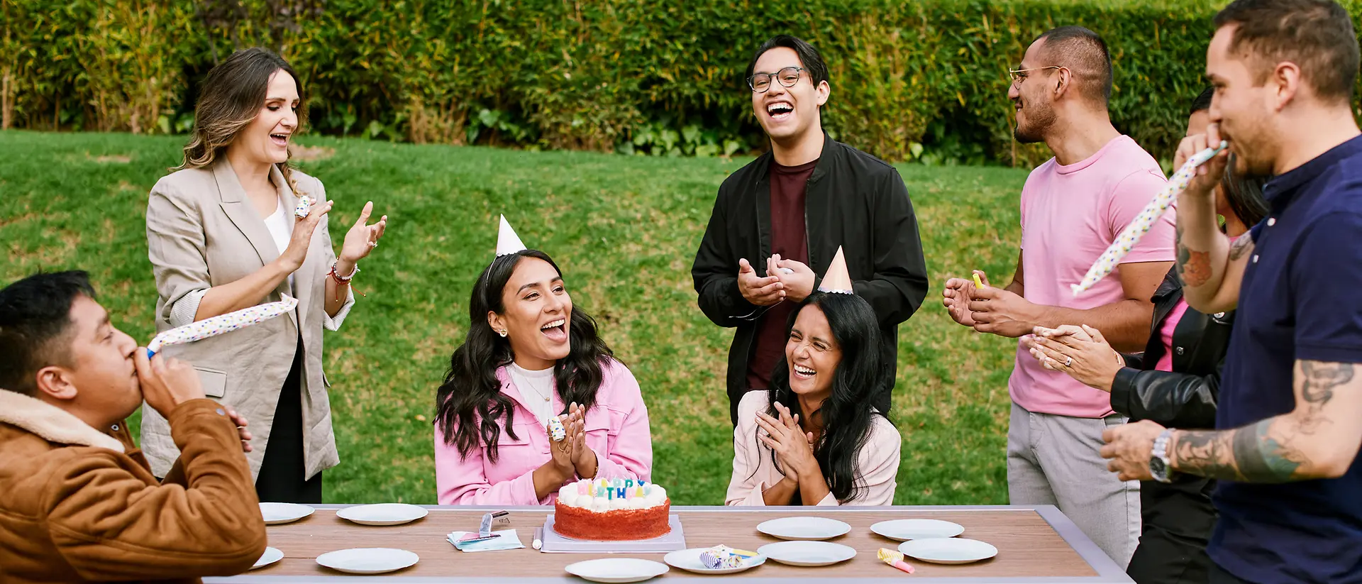 A group of eight colleagues wearing party hats standing in a garden around a desk celebrating the birthday of a woman and laughing together. 