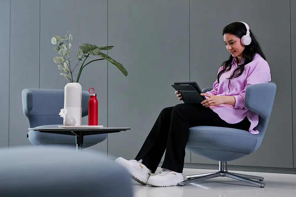 A photo of a woman sitting in a lounge chair focusing on her tablet device while wearing headphones.